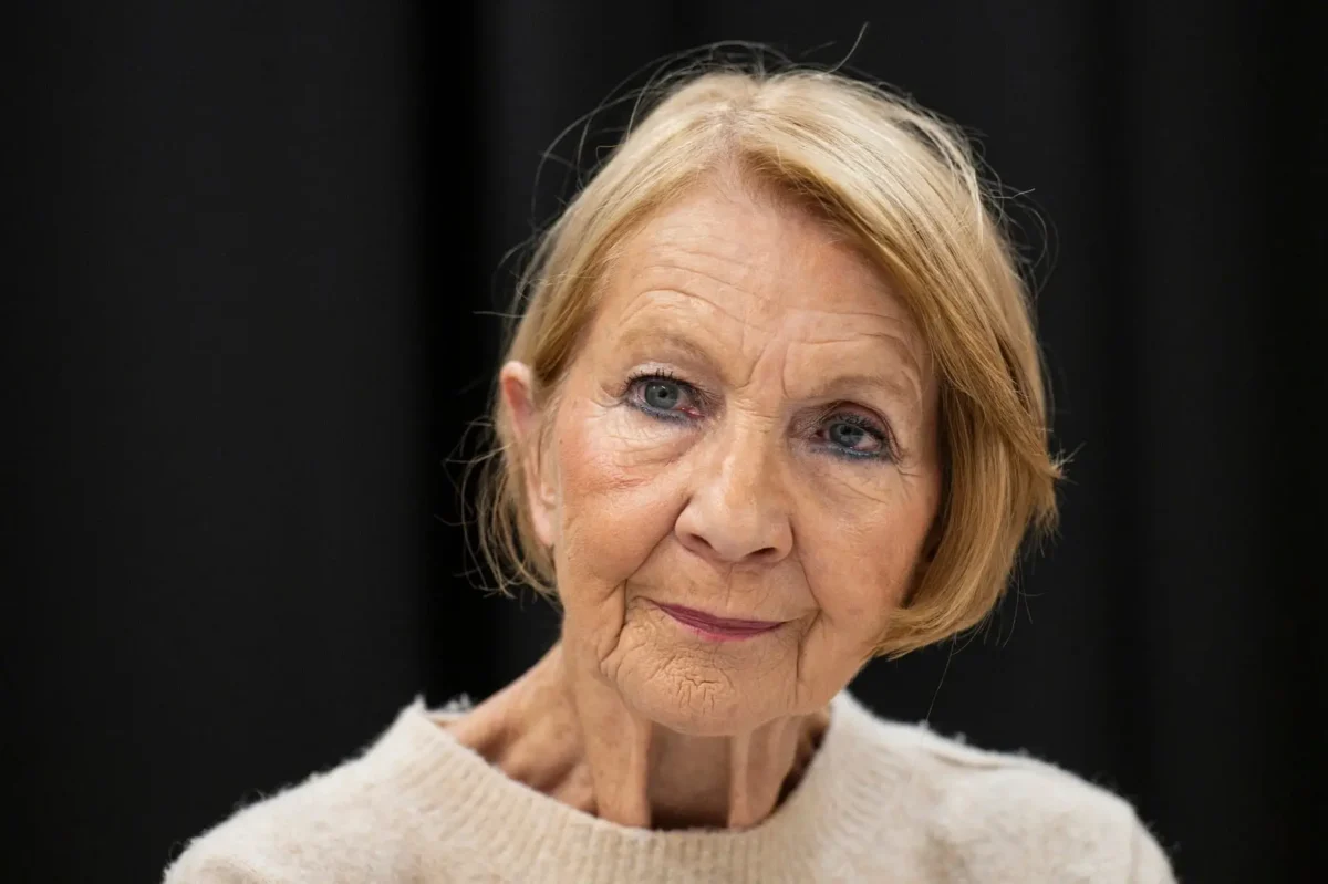 profimedia-1137101811 1 | TVGURU.cz Close-up portrait of an elderly woman with short blonde hair, wearing a light sweater, against a dark background with a gentle smile.