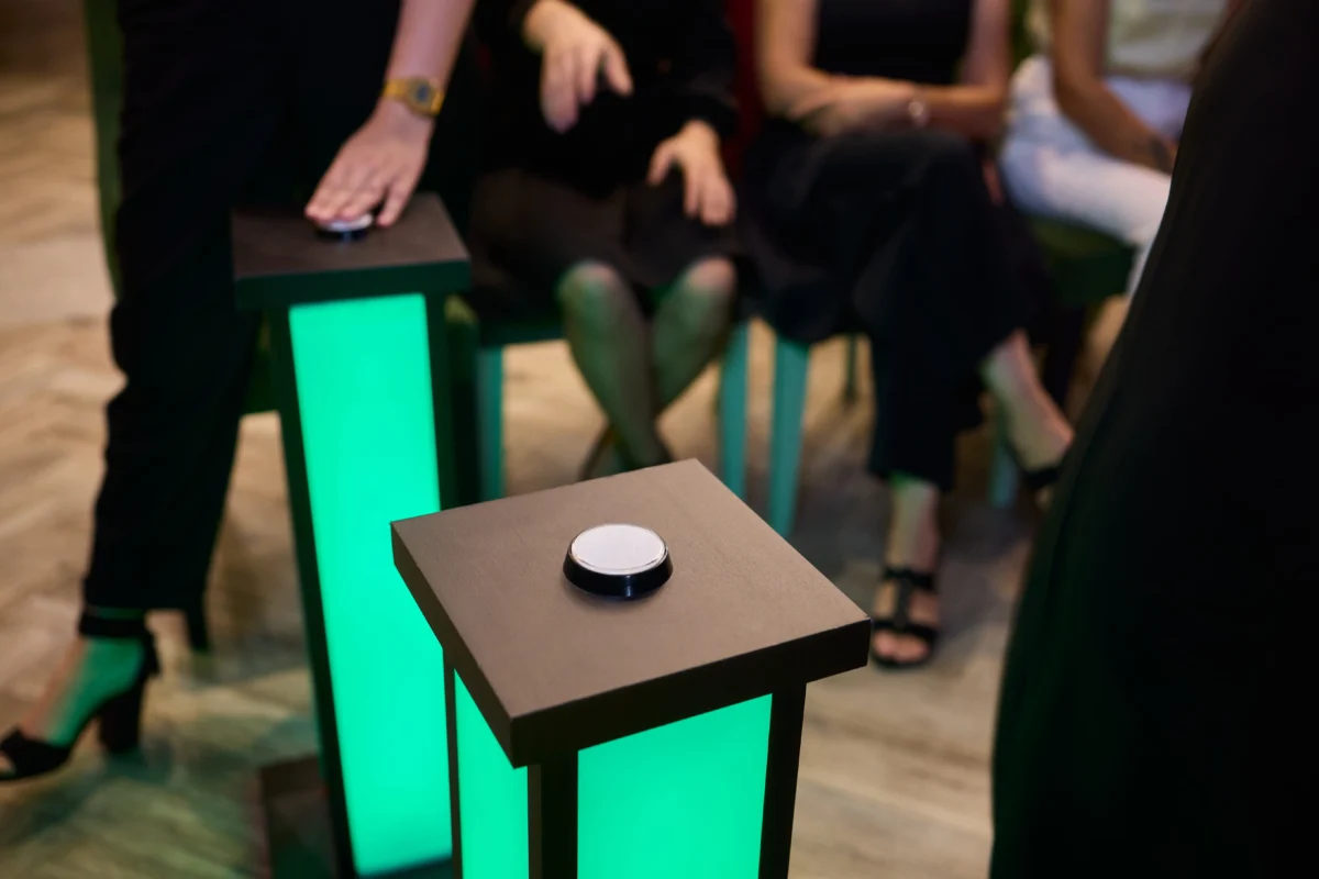 shutterstock_2668148193 1 | TVGURU.cz Close-up of a black square pedestal with a white circular button on top, glowing green panel at an event.