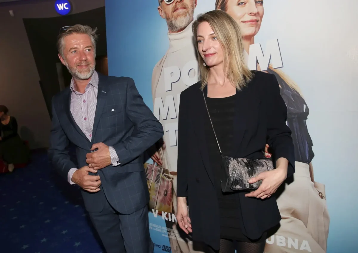 profimedia-0684704542 1 | TVGURU.cz Man in a blue checkered suit smiles while adjusting his sleeve as a woman in black blazer poses beside him in front of a movie poster.