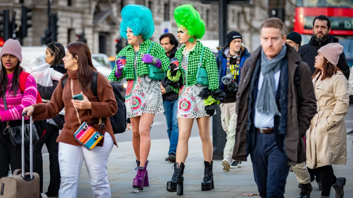 shutterstock_2254954243 1 | TVGURU.cz Two people in neon green wigs and matching houndstooth outfits walk with drinks among a crowded street scene, others around them.