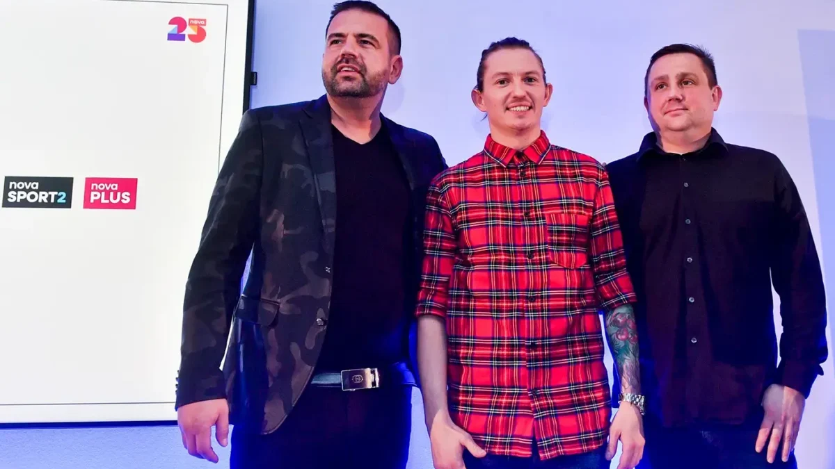 Tři muži stojí na podiu a za nimi na stěně visí promítací obrazovka