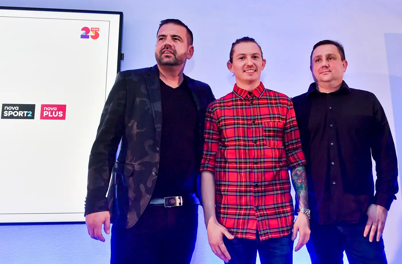 Tři muži stojí na podiu a za nimi na stěně visí promítací obrazovka