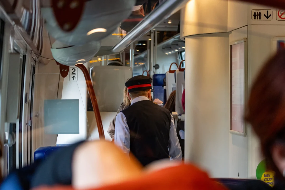 shutterstock_2391761689 1 | TVGURU.cz Crowded train interior with passengers standing and seats visible, a conductor in a dark vest and hat facing away from the camera.