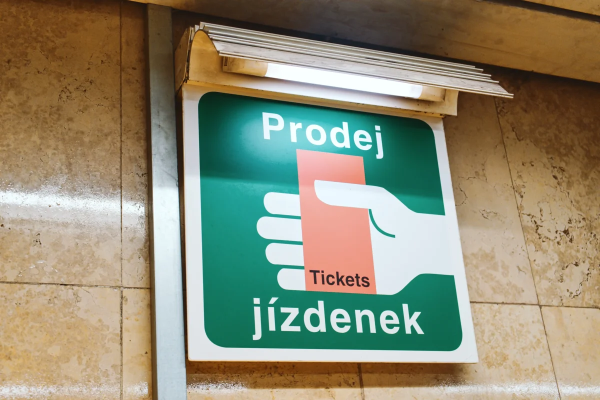 shutterstock_2595315447 1 | TVGURU.cz Green sign reading'Prodej Jízdenek' (ticket sales) mounted on a beige marble wall, with a stack of papers above it.
