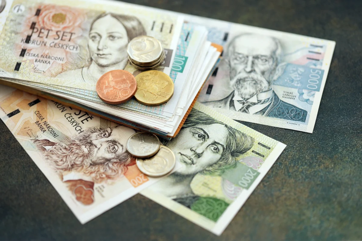 shutterstock_2816390743 1 | TVGURU.cz Pile of assorted Czech banknotes and coins spread on a dark surface, showing various denominations including 1000 Kč notes and coins.