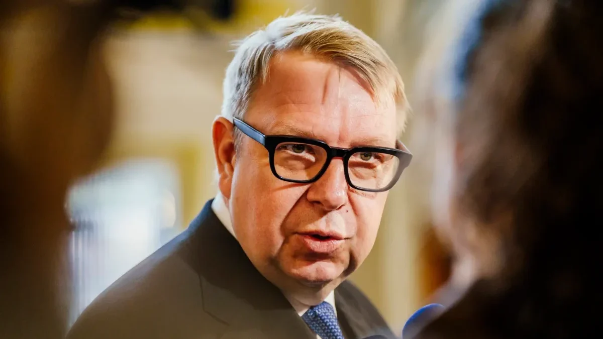 Man in a dark suit and blue tie wearing black glasses, speaking in a blurred indoor setting.