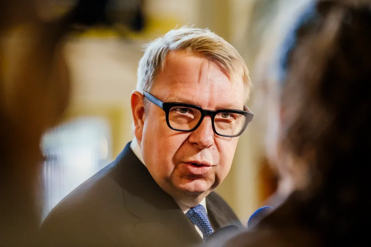 Man in a dark suit and blue tie wearing black glasses, speaking in a blurred indoor setting.