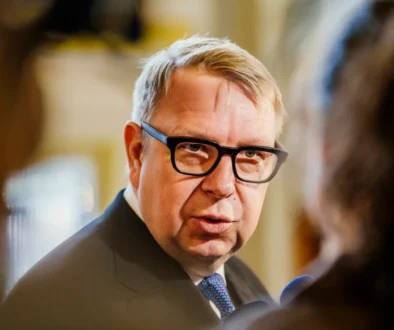 Man in a dark suit and blue tie wearing black glasses, speaking in a blurred indoor setting.