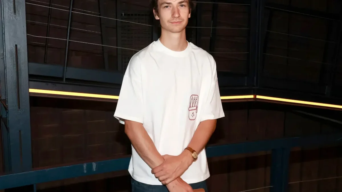 Young man in a white T-shirt and jeans stands in a dim industrial setting with blue metal railings, arms crossed around a wooden object in his hands.