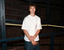 Young man in a white T-shirt and jeans stands in a dim industrial setting with blue metal railings, arms crossed around a wooden object in his hands.