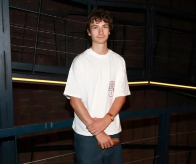 Young man in a white T-shirt and jeans stands in a dim industrial setting with blue metal railings, arms crossed around a wooden object in his hands.