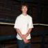 Young man in a white T-shirt and jeans stands in a dim industrial setting with blue metal railings, arms crossed around a wooden object in his hands.