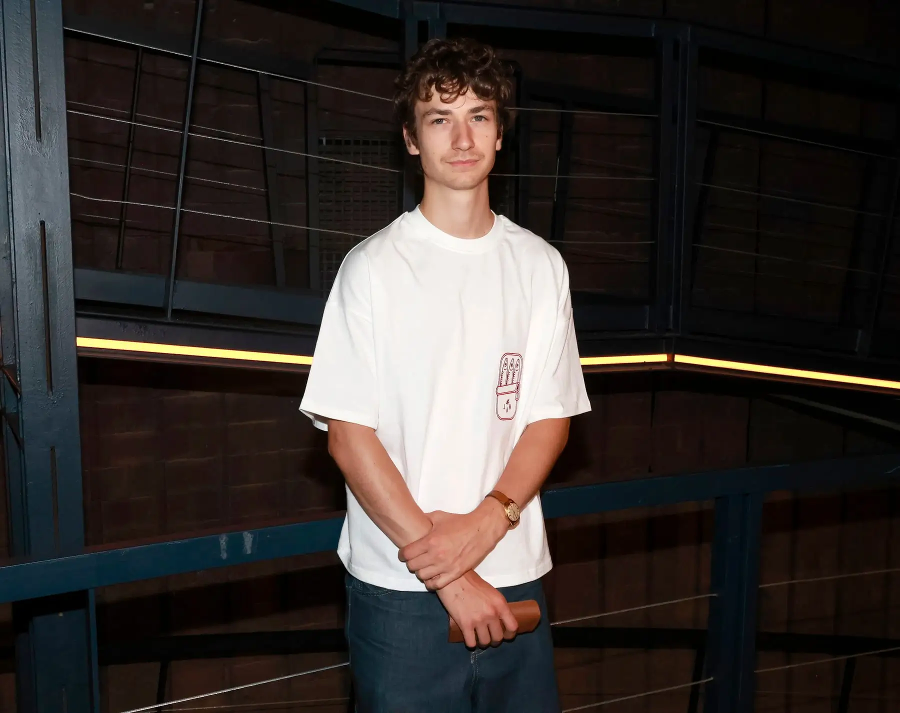Young man in a white T-shirt and jeans stands in a dim industrial setting with blue metal railings, arms crossed around a wooden object in his hands.