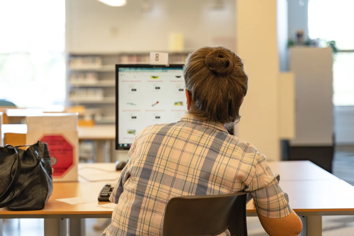 shutterstock_1472280119 1 | TVGURU.cz Person with short gray hair in a blue plaid shirt, seen from behind, sitting at a computer in a bright public workspace.
