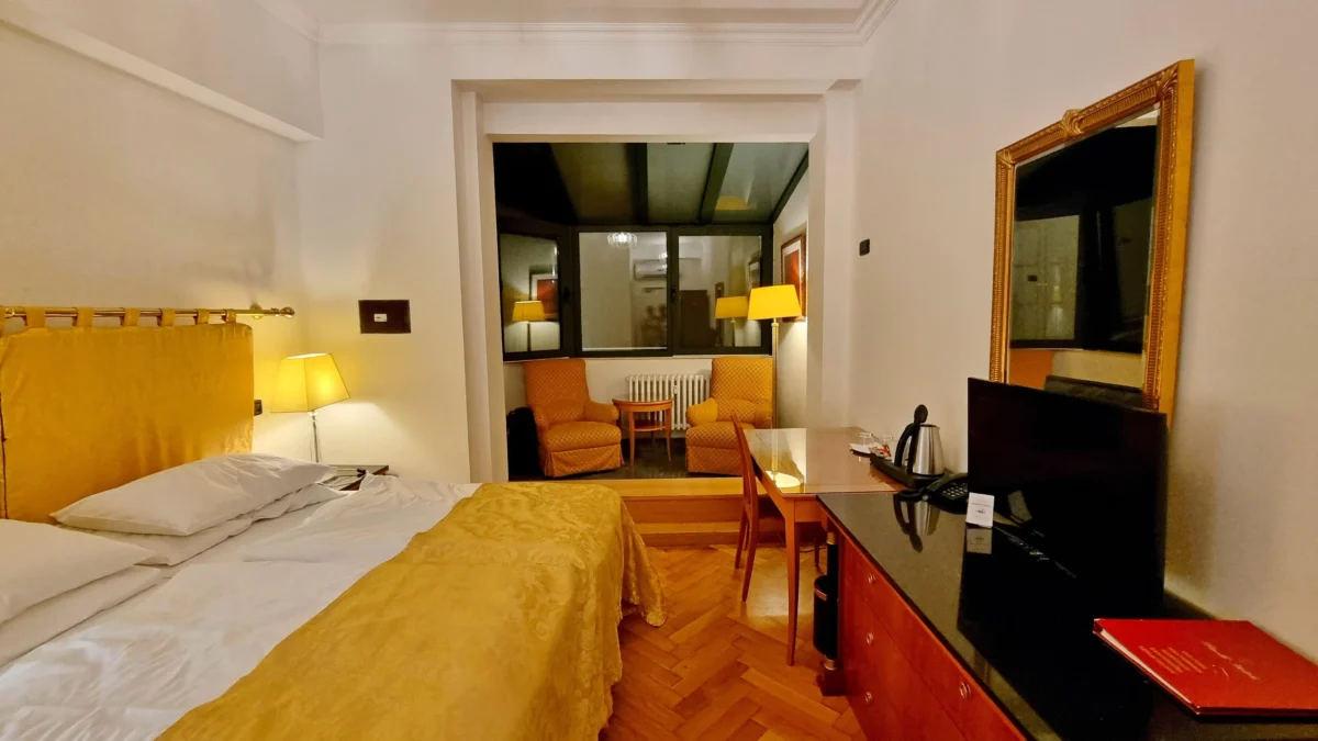 shutterstock_2584922325 1 | TVGURU.cz Hotel room with two large beds in the foreground, gold bedspreads, and a seating area with orange chairs visible through a doorway.