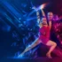Duo of ballroom dancers mid-leap on a neon-lit stage, woman in a pink fringe dress and man in dark attire, with blue and magenta geometric light effects.