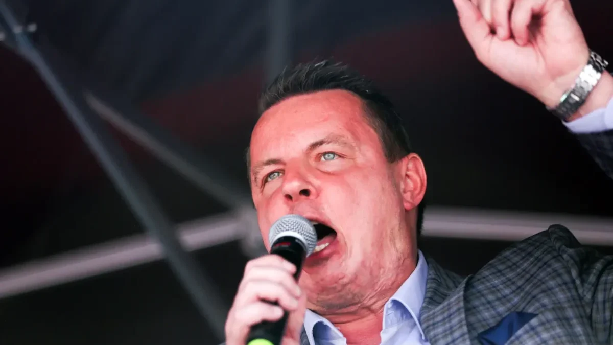 Male singer on stage, microphone in hand, raising one finger as he performs.