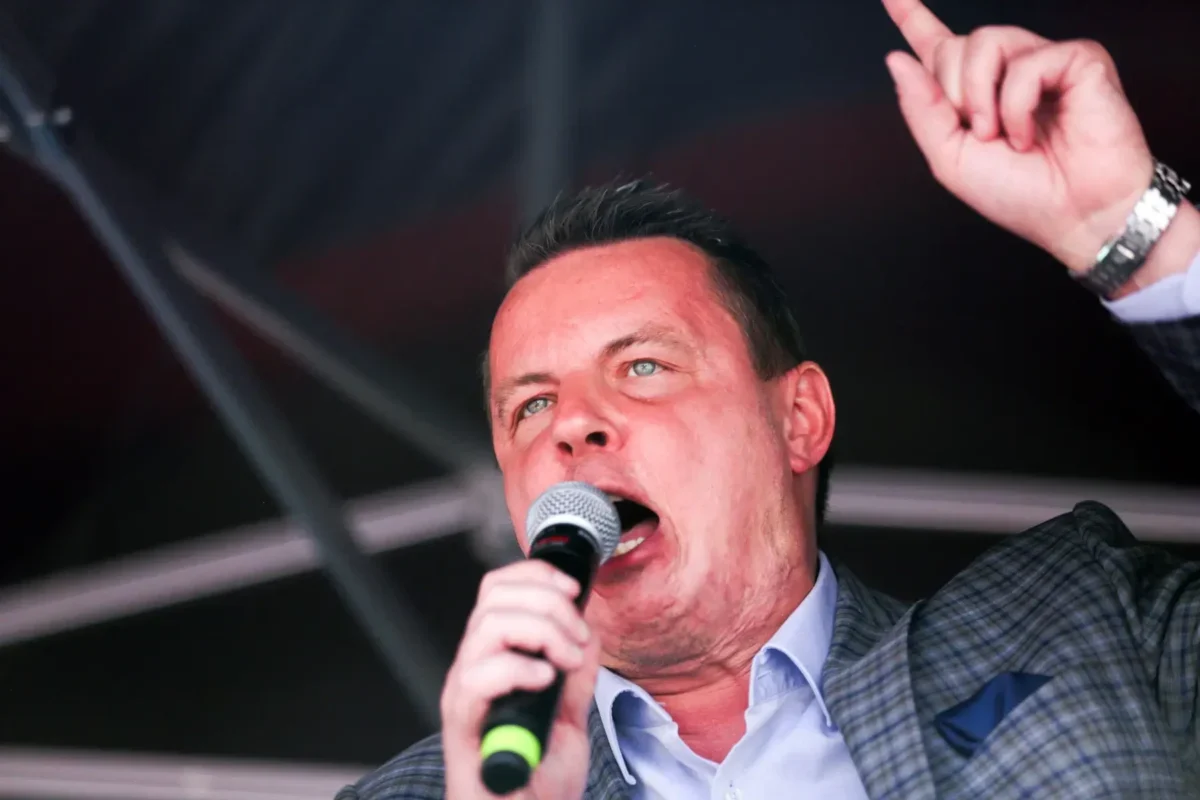 Male singer on stage, microphone in hand, raising one finger as he performs.