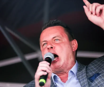 Male singer on stage, microphone in hand, raising one finger as he performs.