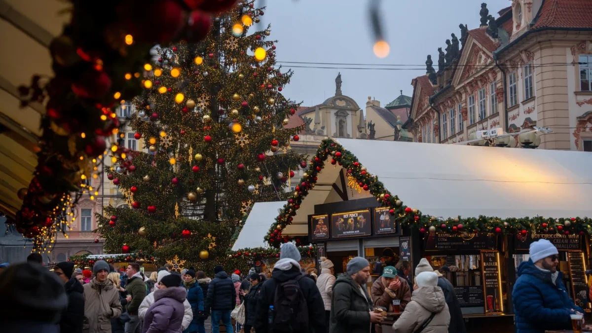 Crowded Christmas market street with a large, decorated tree and festive lights near historic buildings at dusk/evening light framing the scene