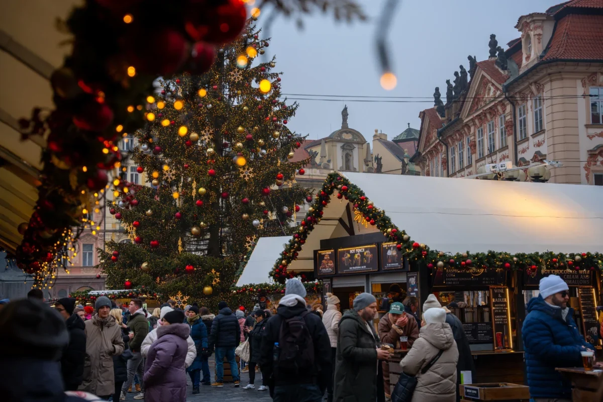 Crowded Christmas market street with a large, decorated tree and festive lights near historic buildings at dusk/evening light framing the scene