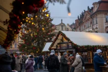 Crowded Christmas market street with a large, decorated tree and festive lights near historic buildings at dusk/evening light framing the scene