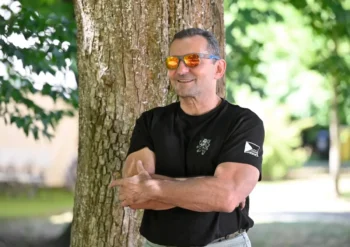 Man in a black t-shirt with arms crossed, leaning against a tree outdoors, wearing orange mirrored sunglasses and smiling in a sunny park setting