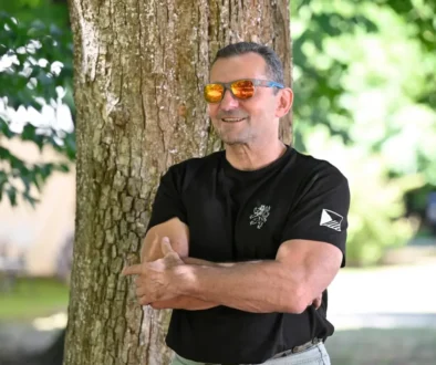 Man in a black t-shirt with arms crossed, leaning against a tree outdoors, wearing orange mirrored sunglasses and smiling in a sunny park setting