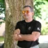 Man in a black t-shirt with arms crossed, leaning against a tree outdoors, wearing orange mirrored sunglasses and smiling in a sunny park setting