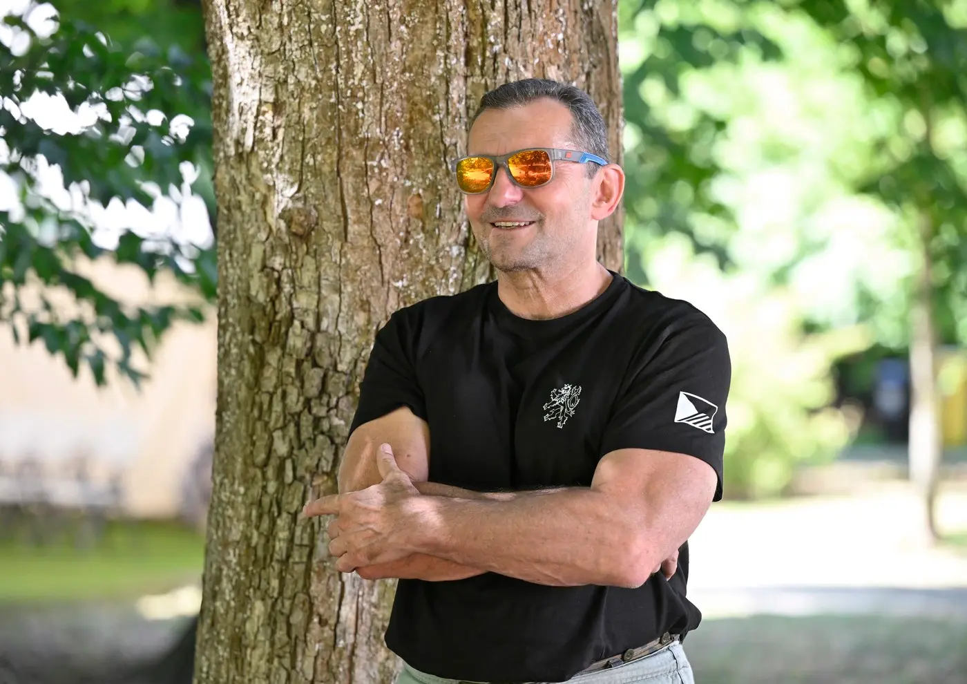 Man in a black t-shirt with arms crossed, leaning against a tree outdoors, wearing orange mirrored sunglasses and smiling in a sunny park setting