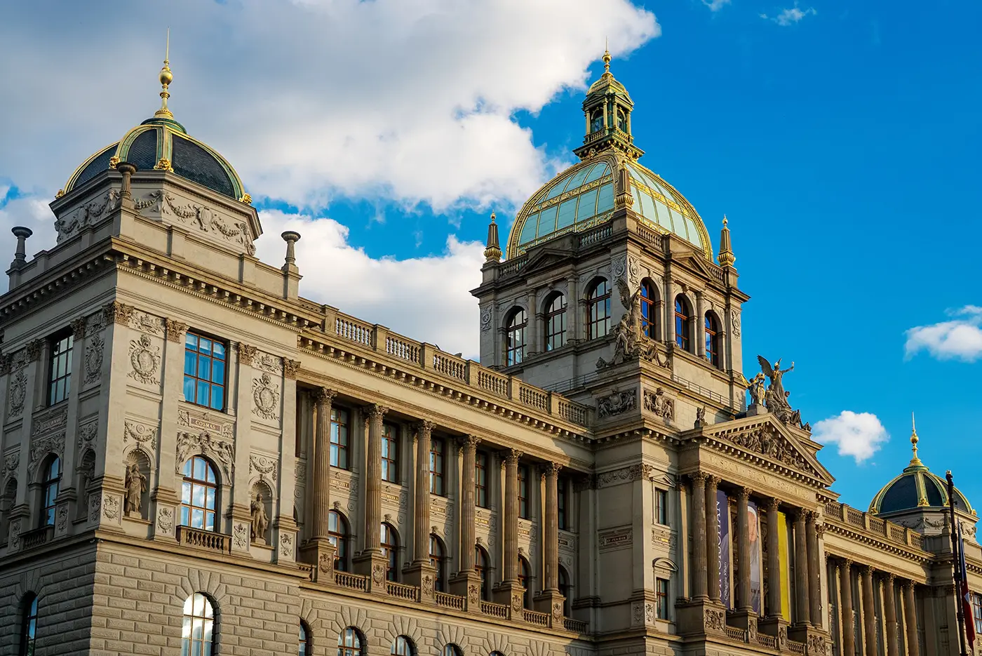 shutterstock_2718161583 | TVGURU.cz Ornate historic building with domed copper roofs and decorative sculptures against a bright blue sky.