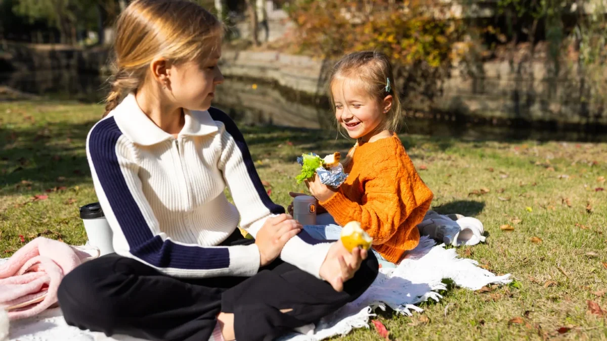 Woman and young girl seated on a blanket enjoying a picnic in a sunny autumn park; they share snacks with a bag and foil-wrapped food nearby.