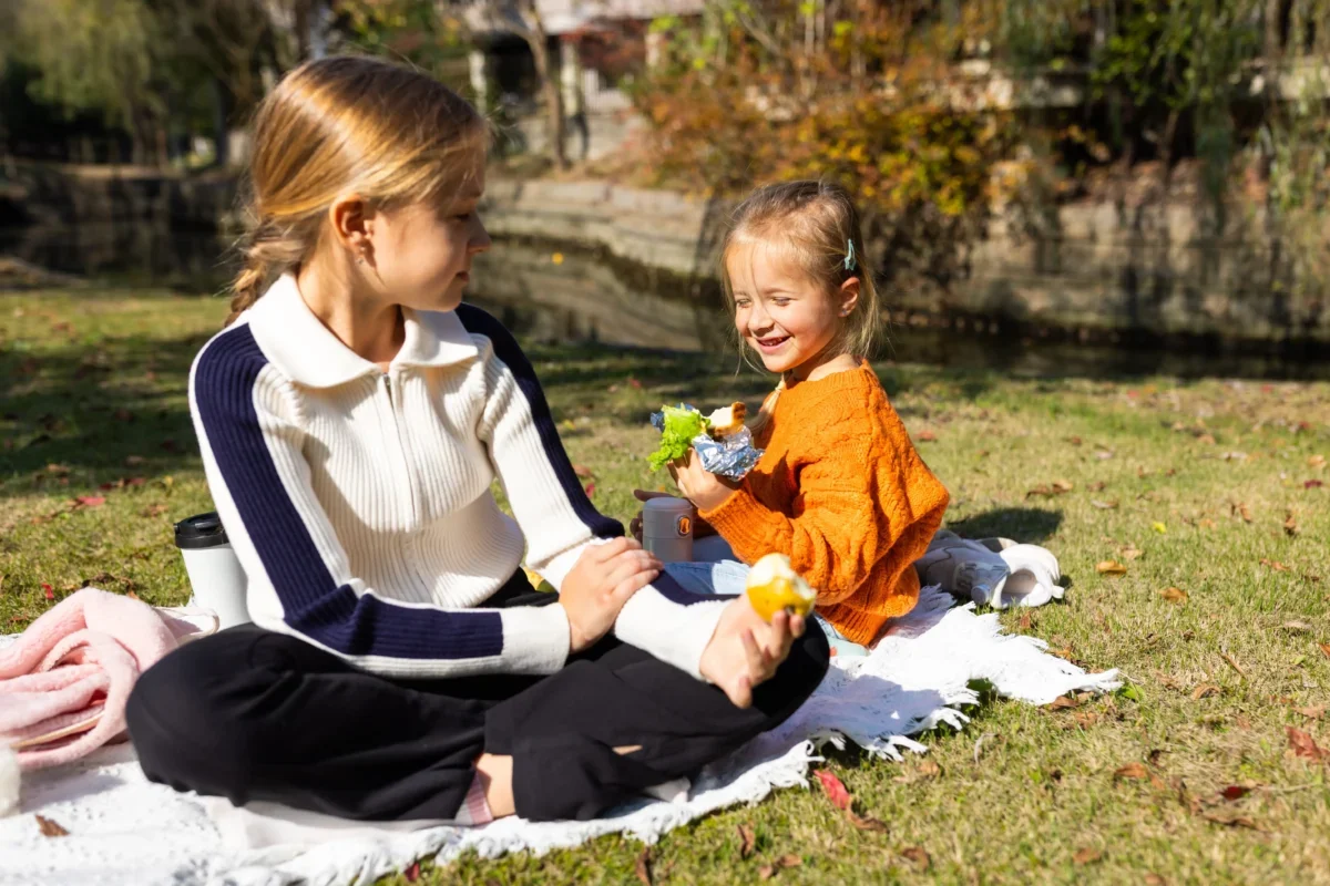 Woman and young girl seated on a blanket enjoying a picnic in a sunny autumn park; they share snacks with a bag and foil-wrapped food nearby.