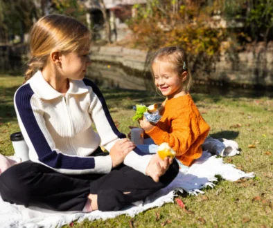 Woman and young girl seated on a blanket enjoying a picnic in a sunny autumn park; they share snacks with a bag and foil-wrapped food nearby.