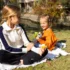 Woman and young girl seated on a blanket enjoying a picnic in a sunny autumn park; they share snacks with a bag and foil-wrapped food nearby.