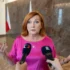 Žena v růžových šatech stojí před novináři a mluví do dvou mikrofonů najednou