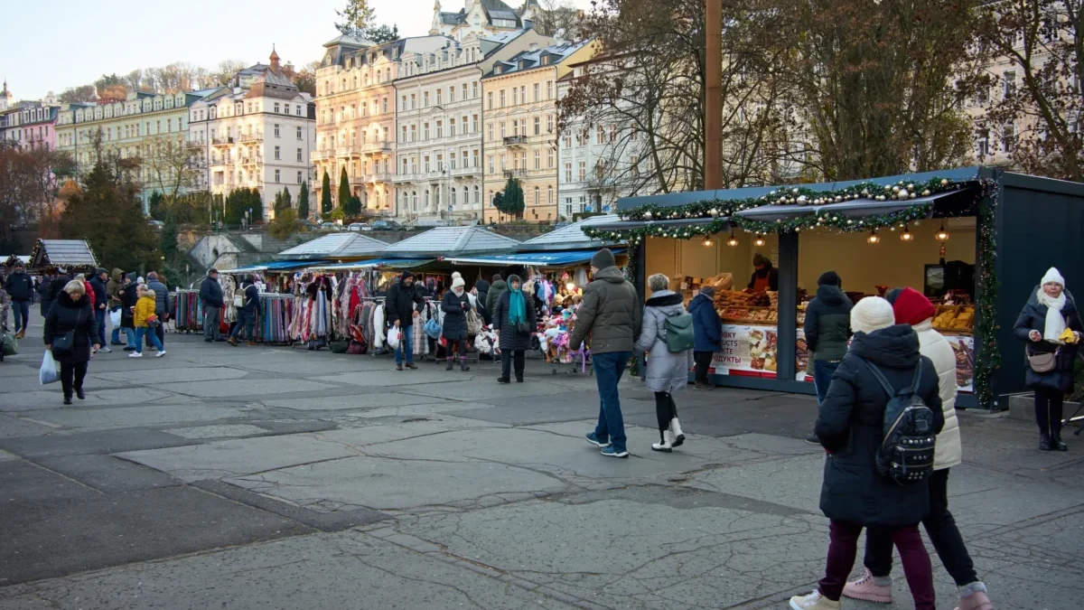 People browse a city market with clothing stalls and festive lights; pastel buildings line the background.