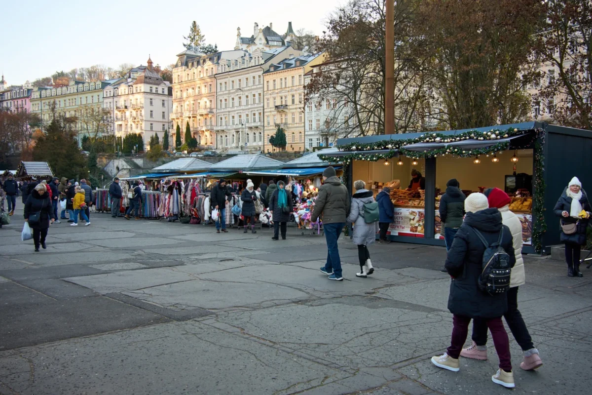 People browse a city market with clothing stalls and festive lights; pastel buildings line the background.