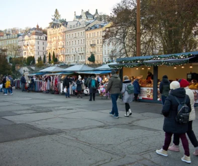 People browse a city market with clothing stalls and festive lights; pastel buildings line the background.