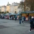 People browse a city market with clothing stalls and festive lights; pastel buildings line the background.