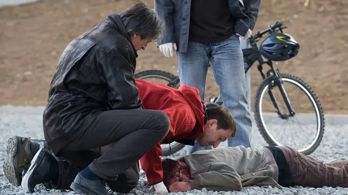 Tři muži se sklánějí nad člověkem ležícím na zemi hlavou dolů