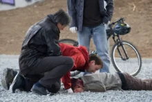 Tři muži se sklánějí nad člověkem ležícím na zemi hlavou dolů