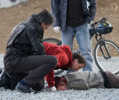 Tři muži se sklánějí nad člověkem ležícím na zemi hlavou dolů