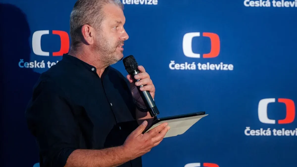 Muž v tmavé košili drží v jedné ruce tablet a papír a ve druhé mikrofon