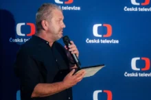 Muž v tmavé košili drží v jedné ruce tablet a papír a ve druhé mikrofon