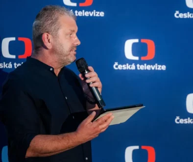 Muž v tmavé košili drží v jedné ruce tablet a papír a ve druhé mikrofon