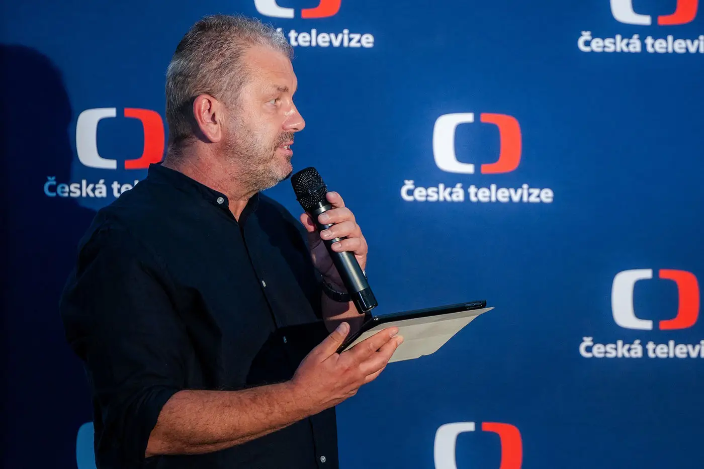 Muž v tmavé košili drží v jedné ruce tablet a papír a ve druhé mikrofon
