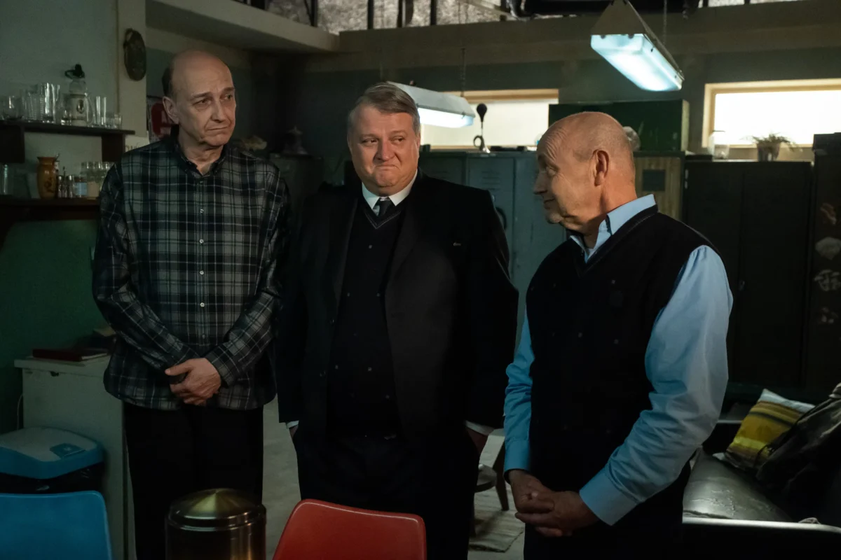 BOZI_PLAN_EPI0007__LUBOS_NOVOTNY_2026_0004 | TVGURU.cz Tři muži vedle sebe stojí v potemnělé místnosti.