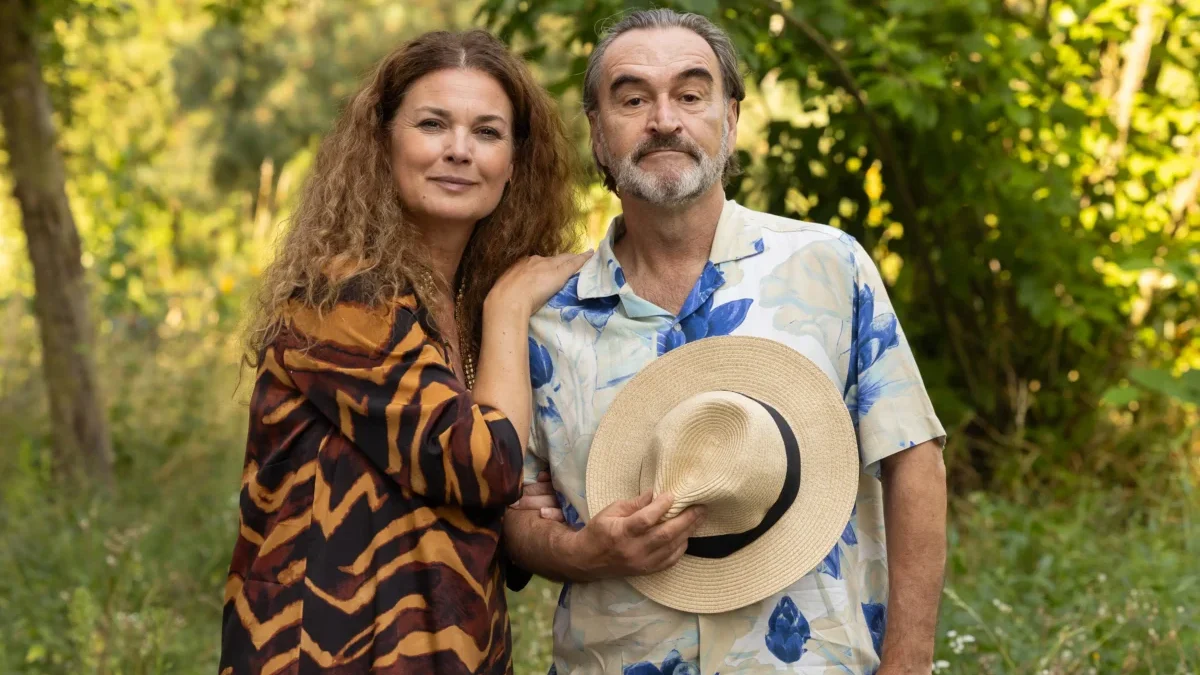 Žena stojí vedle muže a ten si před břichem drží klobouk.