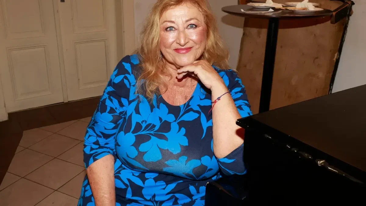 Žena se světlými vlasy v modro-černých šatech sedí na židli a rukou si podpírá bradu